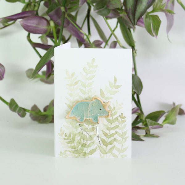 small ceramic and glass aqua blue triceratops dinosaur plant decoration on gift card with watercolour leaves